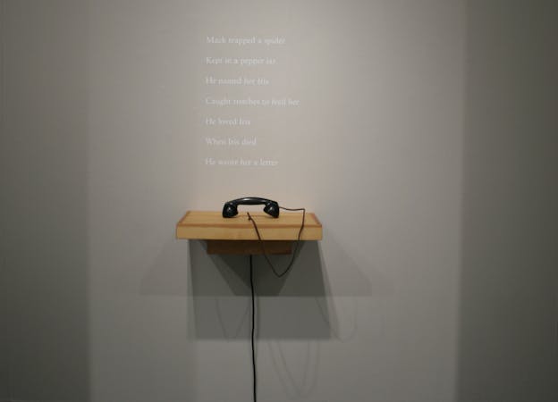 An exhibition photograph of a gray wall with a poem written on it in white text. Beneath the poem there is a wooden shelf with a black telephone on it.