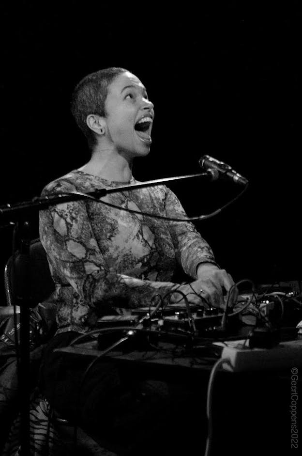 A black and white image of Holland Andrews in front of a mixing board and microphone. Their mouth is wide open, singing, and their eyes are looking upward.