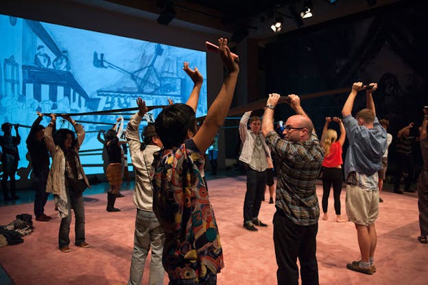 Group of people raising their arms upwards and lifting long boards above their heads on a stage with a blue screen projecting a drawing of people standing in a room.