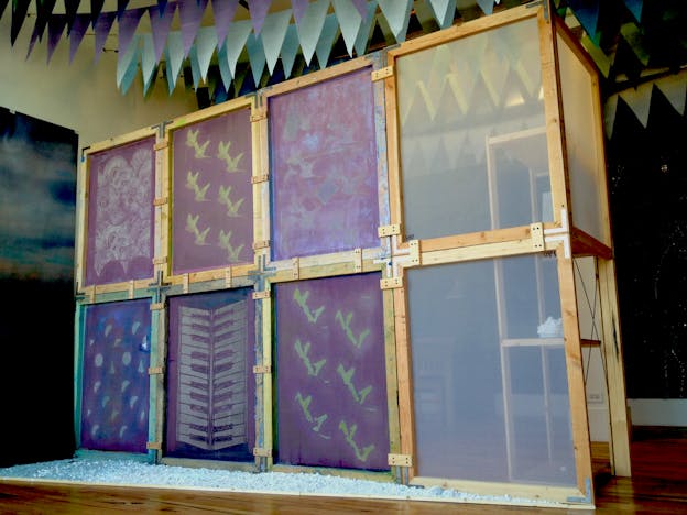 Wooden structure separated into eight squares hosting two transparent screens and six opaque purple screens depicting a flock of birds, a keyboard, crescent shapes, spirals, and other shape patterns.