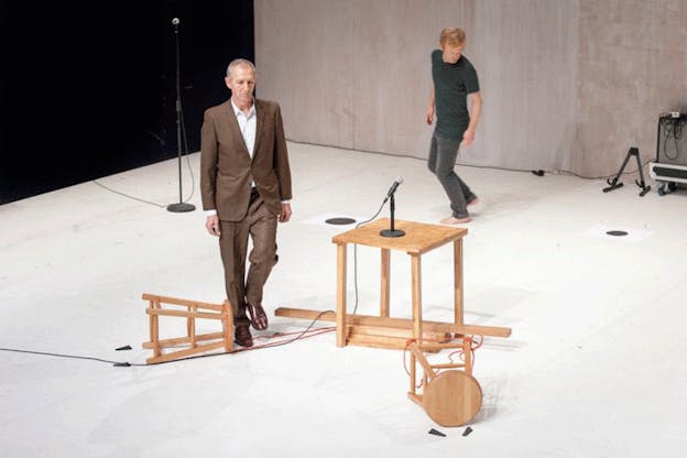 Above view of two performers, one in a brown suit and one in a gray t-shirt and jeans, walking across a white floor with two tipped wooden stools and a small wooden table with a propped microphone. 