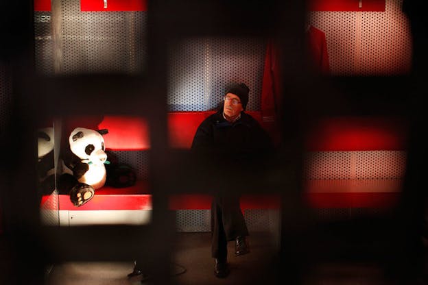 Through the blurred black openings in a structure, a person sits and closes their eyes beside a stuffed panda bear on a lit-up red bench. 
