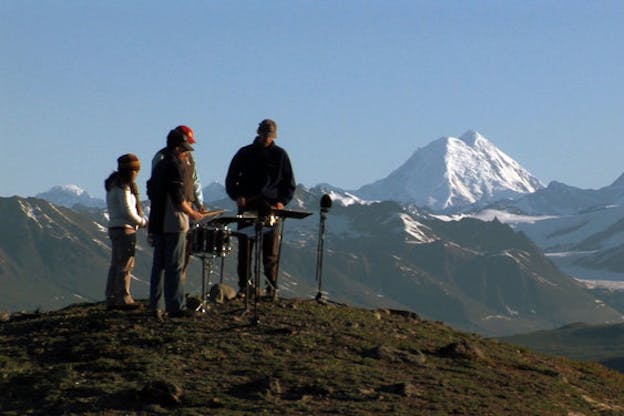 Four figures stand on a grassy mound playing instruments. Snow-topped mountains and a light blue sky are in the background. 