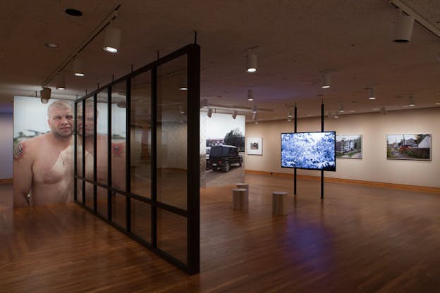 Glass panels separate a photograph of a white man* (person?), his shoulder tattooed with swastikas, from a photograph of a parked jeep and a suspended screen of blue-hued vegetation. On the back wall are framed photographs of residential and high-density housing.
