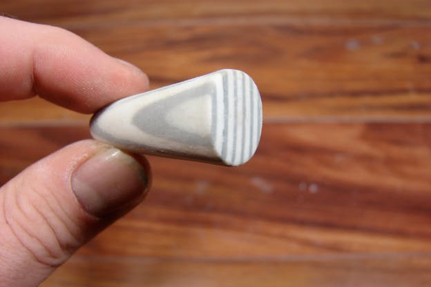 Close-up of two fingers holding a white object resembling a shaven sedimentary stone with gray liesegang rings. 