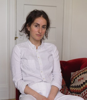 A portrait of Carmen Winant wearing all white, sitting on a red velvet sofa. A multicolored patterned pillow rests next to her.