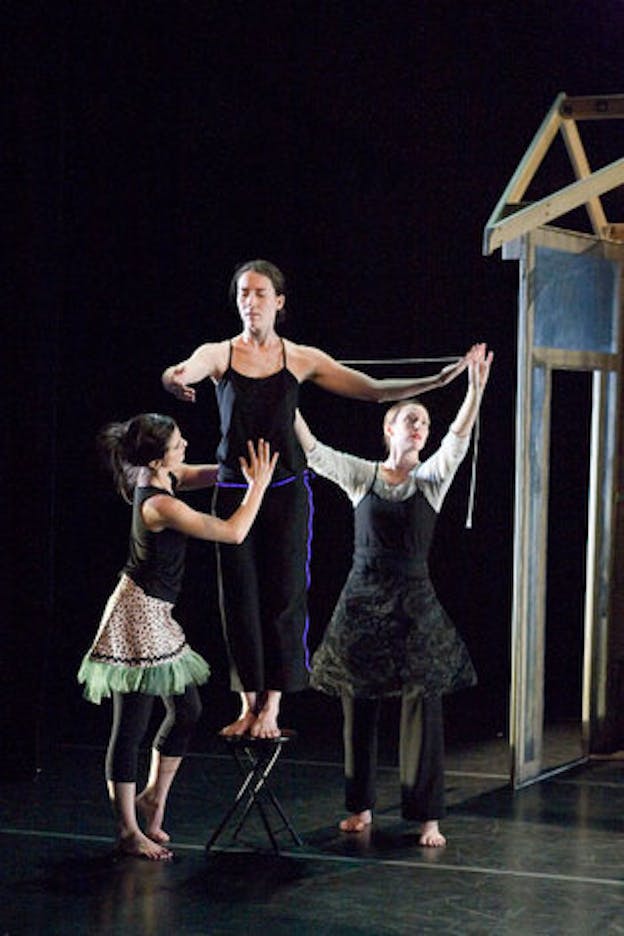 One performer stands on top of a black folding stool with their arms out and two other performers stand on either side of them. One of them touches the front and back of the central performer's abdomen while the other one appears to be measuring the central performer's arm length with a soft tape measure. All three performers are dressed in mismatched outfits. On the right of the image, there is a partial view of a simplified wooden house structure. The performance is staged on a black floor against a black backdrop.