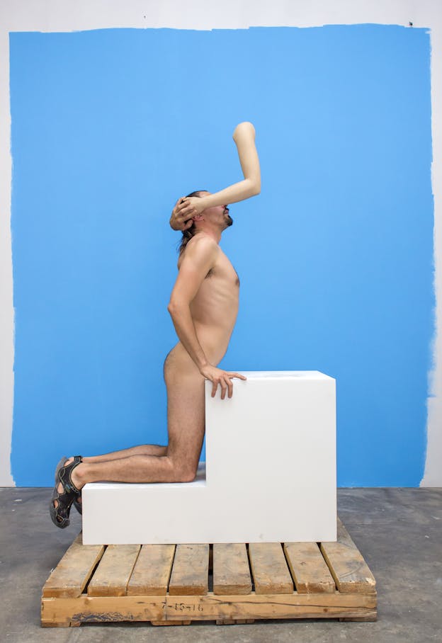 A performer kneels on a white set of two stairs. They are wearing black sandals but are otherwise nude. Their eyes are covered by a disembodied plastic arm which clutches their head. The back wall is light blue and the floor is gray.