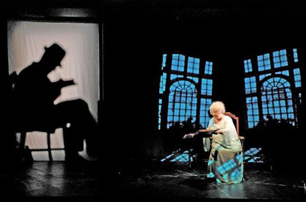 An image of Maleczech performing, dressed in a white loose fitting dress with short, blonde curly hair. She sits on a chair with a large book open in front of her. To her left, there is a larger than life projection of the profile of a man in a fedora sitting on a chair reading a book. In the background, there are inwardly tilted projections of blue lit windows. 
