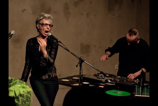 A performer with short gray hair and black framed glasses performing behind a microphone. Behind them further from the viewer a person stands behind a DJ booth. 