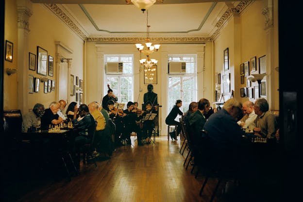 A performance still of a string orchestra in the corner of an ornate room. Around the orchestra, numerous couples are engaged in chess games. The room has numerous framed images on the wall and ornate borders on the ceiling. On the far wall, there are two large windows and a bust sculpture between them. 