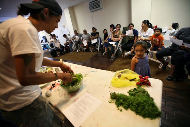 A little girl looks at a person in makeshift stand building a salad. Greenery, radishes in a yellow plastic bag and a paper sit on one side and on the other various spices. Behind them aligned looking towards the stand sit people on white plastic chairs. 
