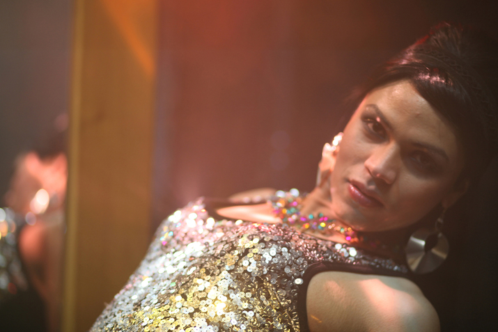 Close-up of a performer in a silver sequin top and dangly silver earrings bending slightly backwards in soft rosy lighting.