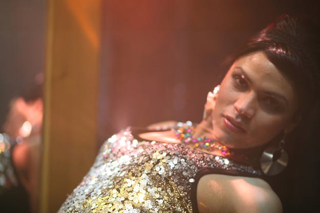 Close-up of a performer in a silver sequin top and dangly silver earrings bending slightly backwards in soft rosy lighting.