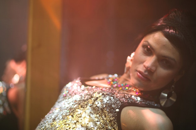 Close-up of a performer in a silver sequin top and dangly silver earrings bending slightly backwards in soft rosy lighting.