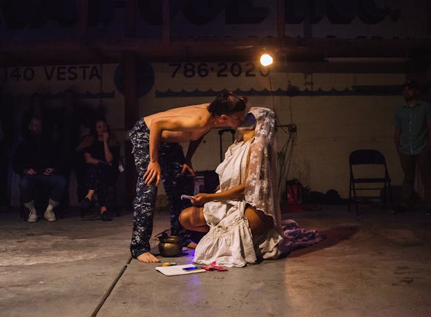 Two performers are situated in the middle of a dimly lit room, kissing each other. One performer crouches, dressed in white lace and the other wears black patterned pants and bends over to reach them. On the floor there are some bits of purple fabric, paper, a cellphone, and a copper pot. Audience members sit against a wall behind them which reads 