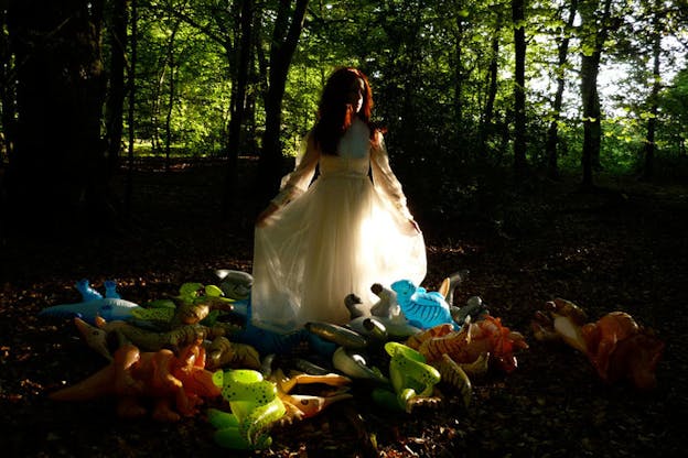 Walsh performs, standing in a white gown and red wig amidst a pile of inflatable multicolored dinosaurs in a dark forest. 