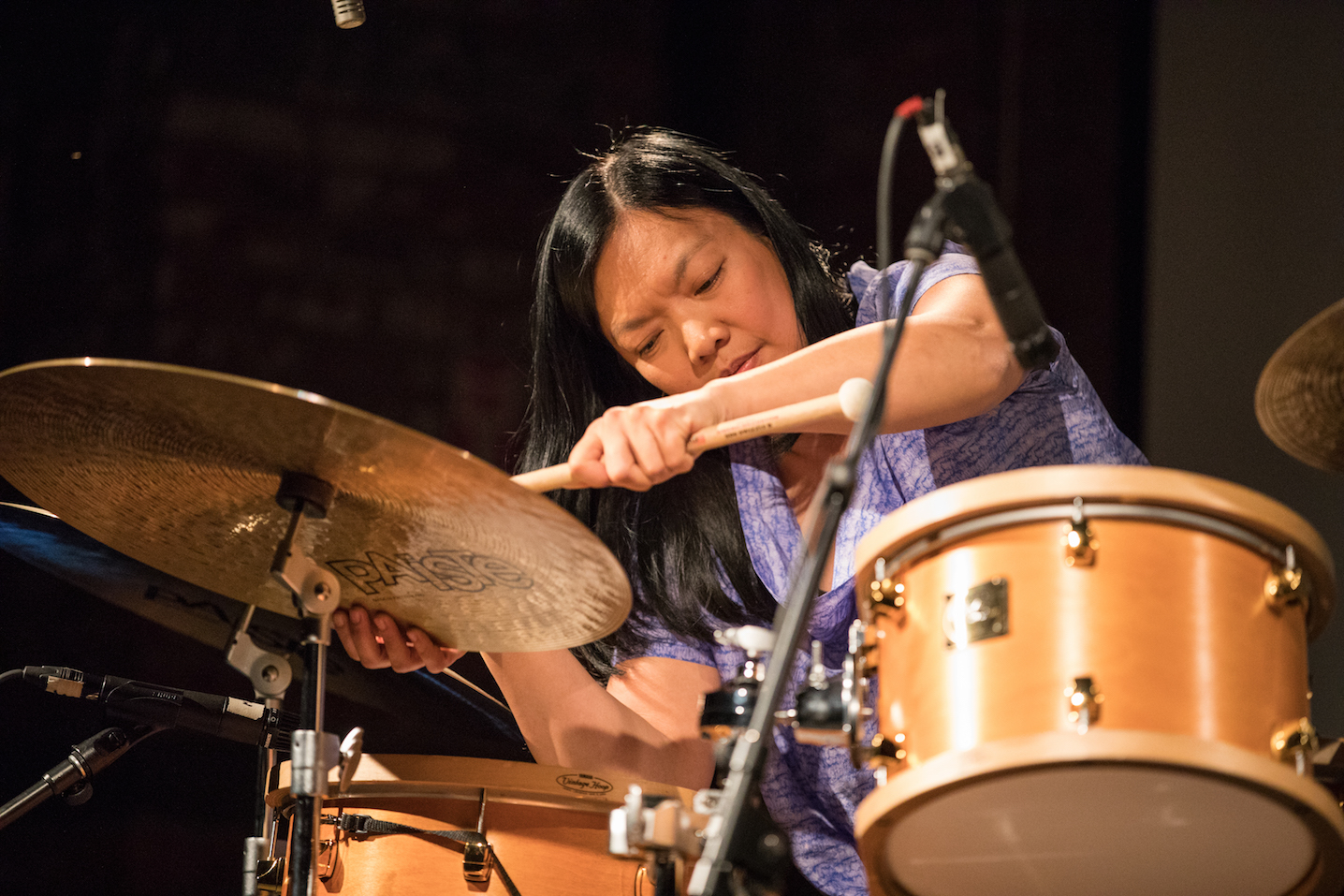 A photograph of Susie Ibarra performing with an orange drumset. She wears a blue patterned shirt and leans over the cymbol, tapping it with a drumstick. The background is blurred black. 