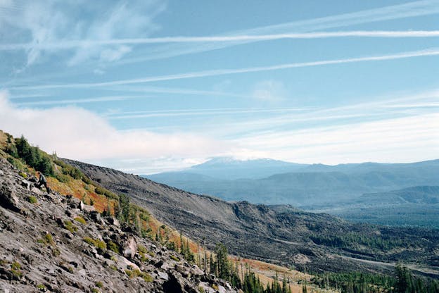 A performance still of the side of a mountain. The forefront is covered by many rocks. Just beyond that, there is a thin line of pine trees on red soil. In the background, numerous other mountains are visible. The sky is bright blue with a large cloud on the left and thin stripes of clouds criss-crossed elsewhere. 