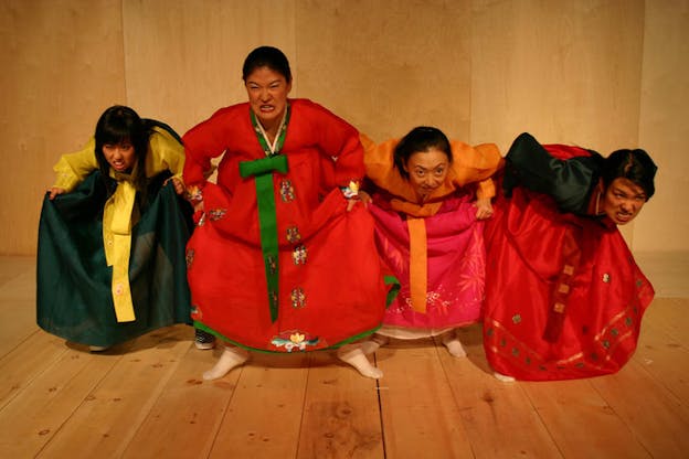 Four people dressed in traditional Korean wear bend over in a squat position with angry facial expressions. The set has wood floors and walls.