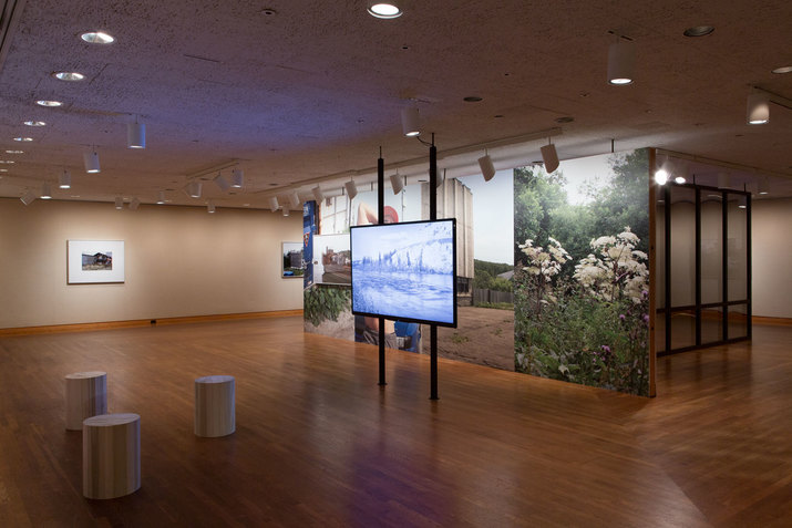 Three wooden stools face a light blue screen mounted in front of four panel screens, one screen visibly displaying an image of white and purple flowers.