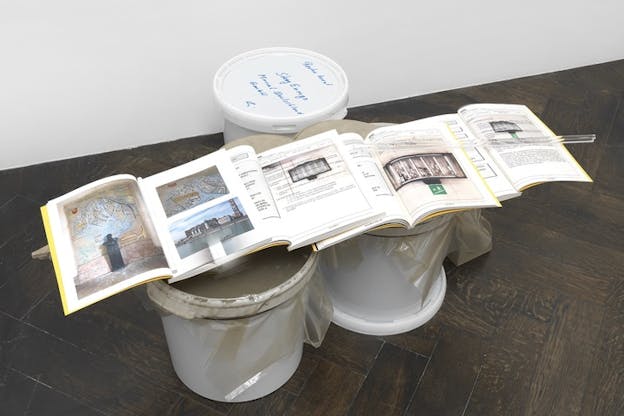 Four open books on top of each other supported by two upside down buckets. A third bucket behind them with the cap on has writing on it.