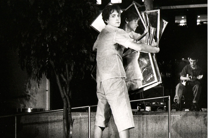 A black and white image shows a performer wearing a t-shirt and capri pants holding several square mirrors in front of them. They turn their head back, away from the mirrors and towards the camera so that the side of their head is reflected in the mirrors. In the background, there is a small tree rows of theater audience chairs and a person sitting in one of the chairs in the front and looking down.