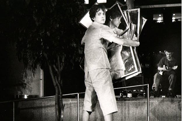 A black and white image shows a performer wearing a t-shirt and capri pants holding several square mirrors in front of them. They turn their head back, away from the mirrors and towards the camera so that the side of their head is reflected in the mirrors. In the background, there is a small tree rows of theater audience chairs and a person sitting in one of the chairs in the front and looking down.