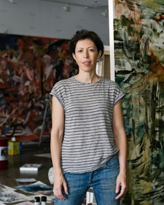 Portrait of Cecily Brown with brown curly short hair, dressed in a black and white striped shirt and jeans. Behind them abstract paintings with artist materials on the floor. 
