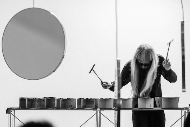 Keiji Haino hunches over a long percussion instrument holding two hammer-like mallets. He wears black clothing. On the left is a large hanging circle.