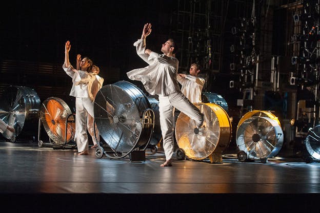 Performers wearing white tunic clothing raise one leg in low arabesque and look up towards their 90-degree angle bent arm. On the dark stage are rows of metal fans in varying sizes.