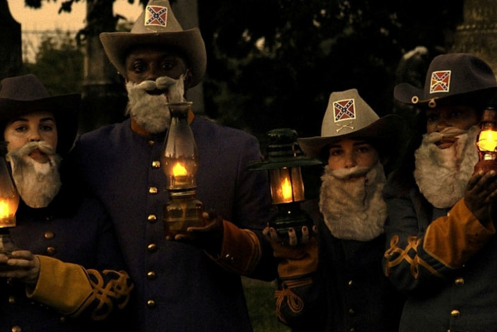 A photograph of three performers wearing matching hats, beards, and holding lanterns. All performers look directly at the camera. The background is a blurred outdoor scene.