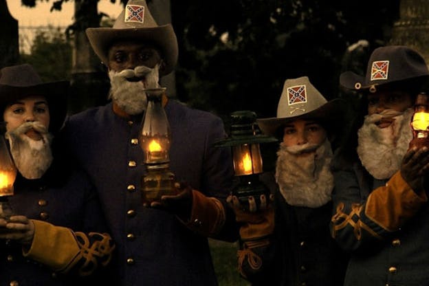 A photograph of three performers wearing matching hats, beards, and holding lanterns. All performers look directly at the camera. The background is a blurred outdoor scene.