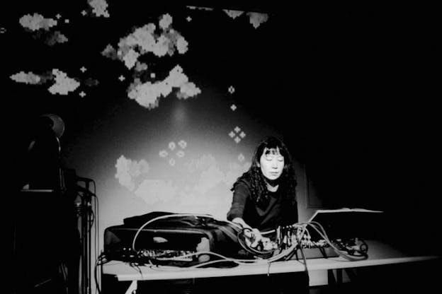 A black and white photograph of Ikue Mori working with electronic music sets on a table. A projection of pixelated patterns is shown in the background.