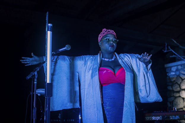 A performer dressed in a pink-blue dress, pink headpiece and a kimono style cardigan stands on blue lighted stage behind a microphone.