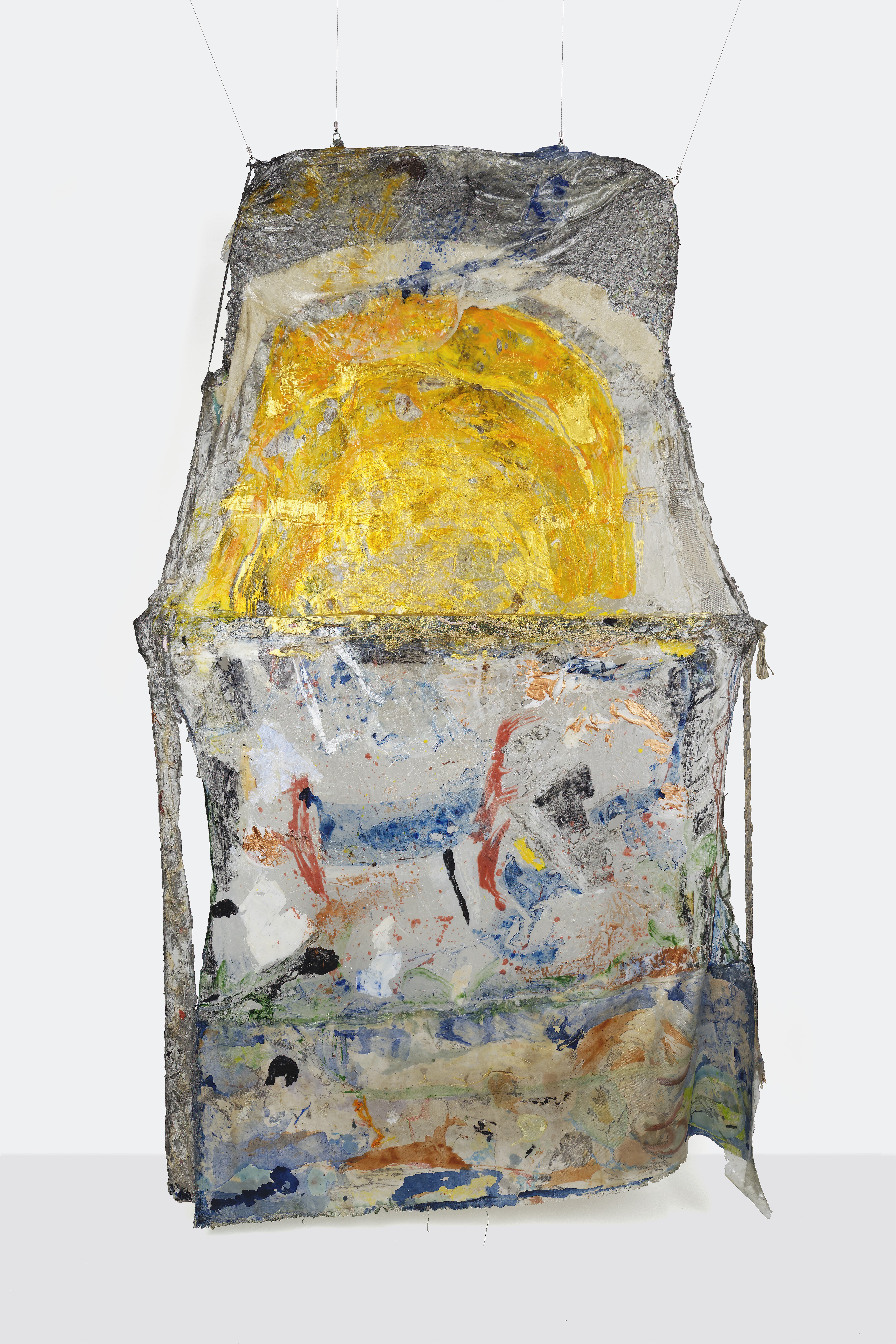 A hanging sculpture made of stretched gray material over a straight-edged frame. The top half of the mostly rectangular piece has a trapezoidal shape, the two outer edges pointing inward. The top half is painted with a bright yellow semicircle with an opaque off white stripe painted above. The material above this stripe is crinkled aluminum with a thin layer of clear plastic draped over, flecked with yellow and dark blue paint. The bottom half below the trapezoidal portion is similarly composed of a mix of stretched aluminum foil, plastic, and fabric, smeared with paint of various colors including red, green, dark blue, black, white, and metallic bronze.  