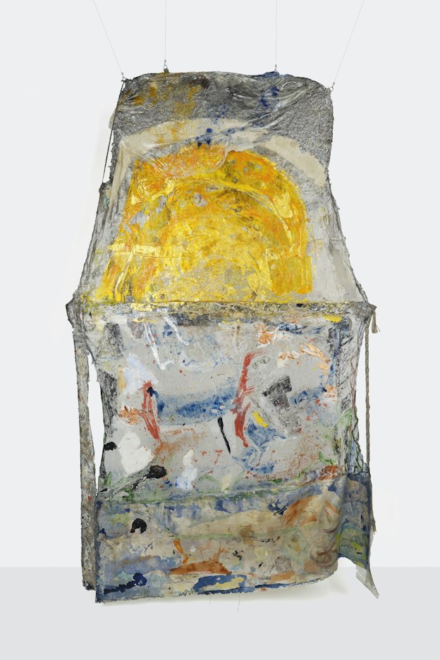 A hanging sculpture made of stretched gray material over a straight-edged frame. The top half of the mostly rectangular piece has a trapezoidal shape, the two outer edges pointing inward. The top half is painted with a bright yellow semicircle with an opaque off white stripe painted above. The material above this stripe is crinkled aluminum with a thin layer of clear plastic draped over, flecked with yellow and dark blue paint. The bottom half below the trapezoidal portion is similarly composed of a mix of stretched aluminum foil, plastic, and fabric, smeared with paint of various colors including red, green, dark blue, black, white, and metallic bronze.  