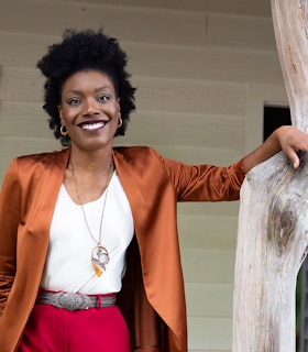 A portrait of Jenn Freeman leaning with her left hand against a wooden column, smiling towards the camera, wearing a metallic burnt copper colored blazer over a white shirt with red pants.