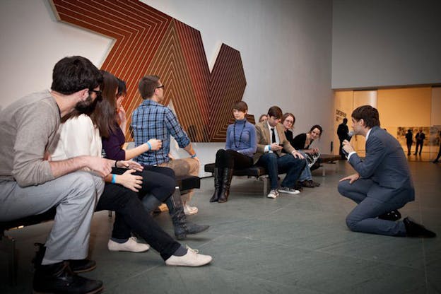 Two bench rows of people watch as two persons face off in a staring contest moderated by a suited person kneeling and holding a microphone. 