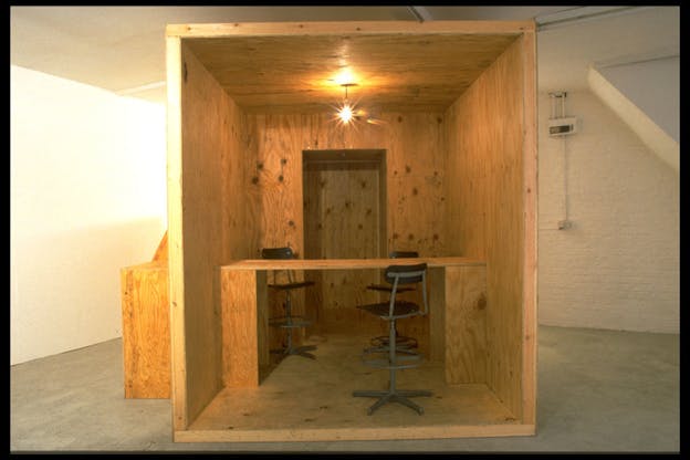 A big wooden cube inside a white brick room has a lamp shining inside it and three roller chairs surrounding a makeshift wooden bar table.