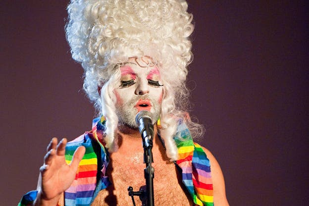 A bare-chested performer wearing rainbow cloth around their shoulders, a curly platinum beehive wig wreathed with pearls, and thick-white face paint, closes their pink and gold fake eyelashed eyes and sings into a microphone while raising one palm. 