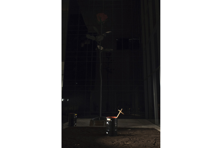 Photograph of a black trash can at night with a wooden cross sticking out of it. Behind it, there is a thin tree. 