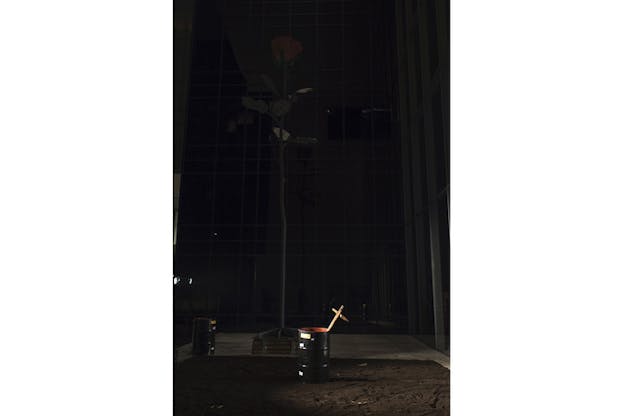 Photograph of a black trash can at night with a wooden cross sticking out of it. Behind it, there is a thin tree. 