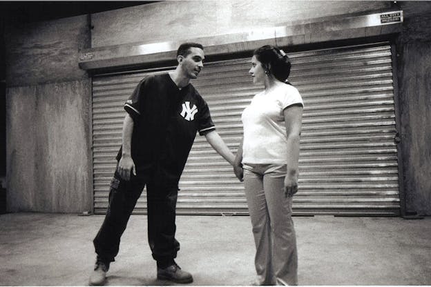 A black and white image of two performers who stand in front of a closed metal gate in the middle of wooden walls. One performer wears dark pants and a New York Yankees shirt and leans towards the other performer to take their hand. The other performer wears a light t-shirt and dark pants and has their long dark hair in an updo. They look hesitant. 
