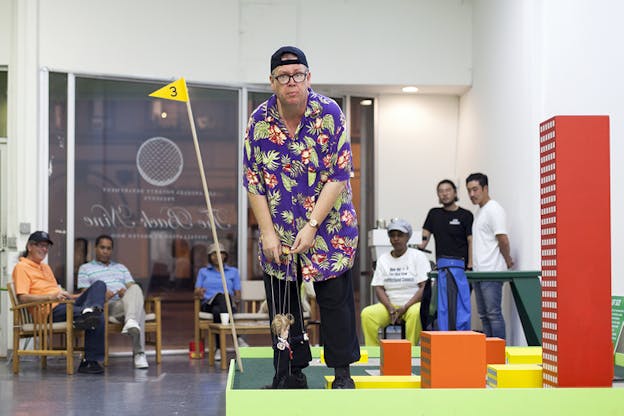 A figure in a purple flowery button up and upside cap holds a doll by the strings on a colorful cities macket. An audience watches from behind.