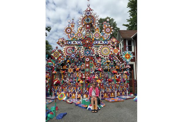 Beglarian sitting and smiling in front of a bright carnival-colored cross painted with abstract shapes and symbols of crescents, wheels, circles, stars, and diamonds. 