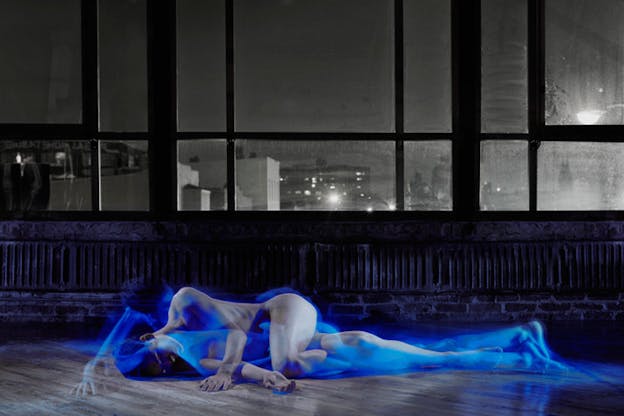 Jonah Bokaer is depicted in black and white as he rests his body on the floor, laying sideways and nude. A transparent, blue hued image of Bokaer laying sideways with his head raised and supported by his hand is overlayed over the black and white image. The floor is wood paneled and the walls behind Bokaer contain large windows that look out onto a cityscape at night.