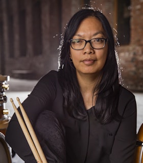 A portrait of Susie Ibarra wearing a black button up shirt and black glasses. She holds a pair of drumsticks. Behind her, there is a red brick wall and the edges of a drum set. 