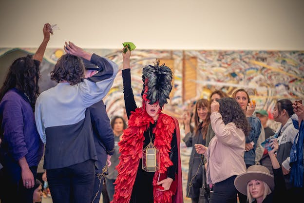 A figure dressed in a coat of red feathers, headpiece of black and red feathers and an empty oil lamp around their neck passes through a crowd with their arm extended upwards holding a green foil.