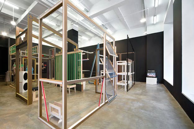 An installation image of numerous wooden structures painted black, green, blue, and red. They form rectangles, triangles, grids, and shelves. On the far right of the room, there is a small television depicting a brightly lit, pink image on it.